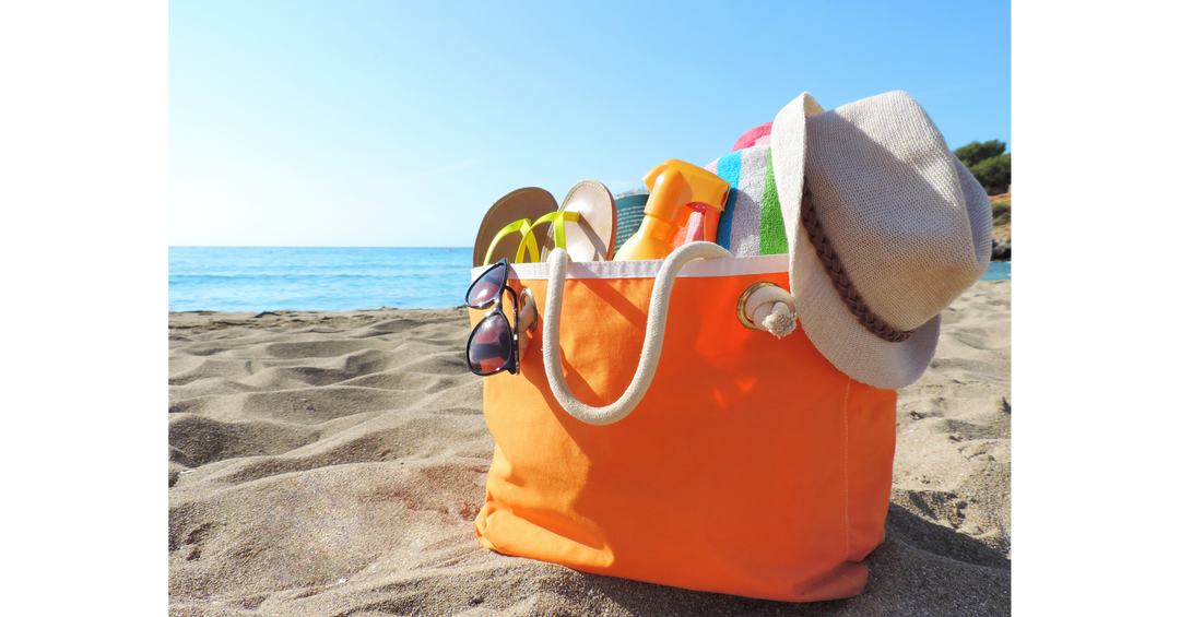 A bright orange beach bag filled with summer essentials like sunscreen, flip-flops, towels, sunglasses, and a sunhat on the sand by the ocean.