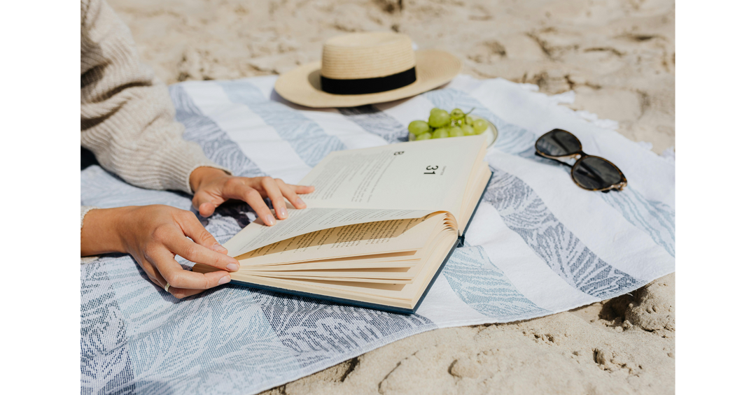 Person reading an open book on a striped beach towel beside a sunhat, sunglasses and grapes during a relaxing day at the beach.