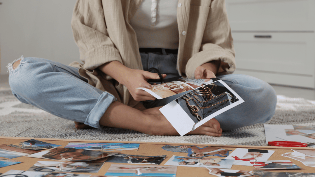 A person sitting cross-legged on the floor cutting printed photos for a vision board.