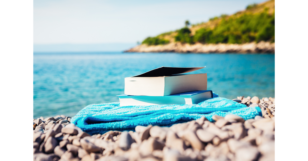 A stack of books resting on a bright blue towel beside the ocean on a rocky beach.