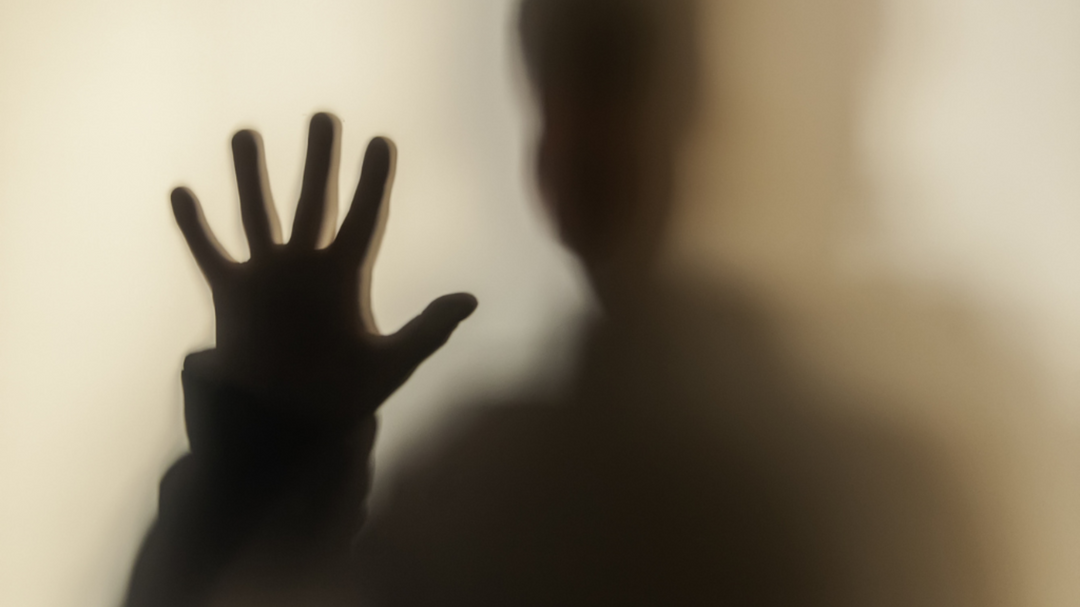 A shadowy silhouette of a person with their hand pressed against frosted glass, creating a suspenseful and eerie atmosphere.