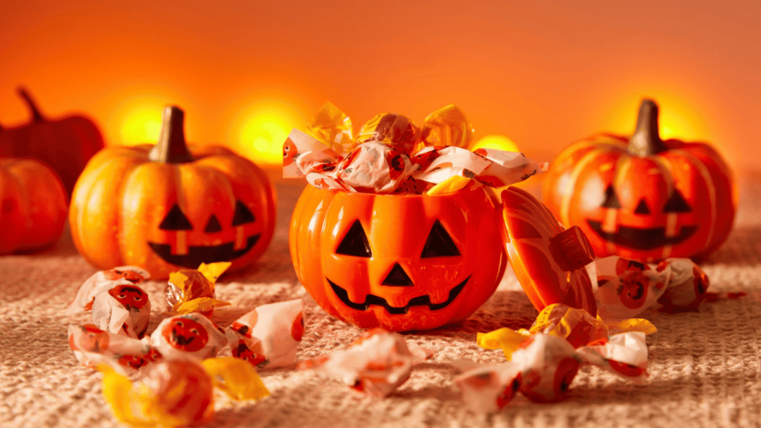 A plastic jack-o'-lantern filled with Halloween candies sits on a cozy surface surrounded by more pumpkins and glowing lights.