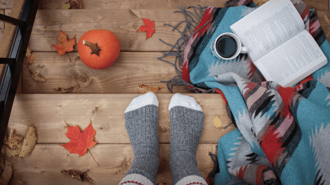 A cozy fall scene with socked feet on a wooden deck, autumn leaves, a pumpkin, a blanket, coffee mug, and an open book.