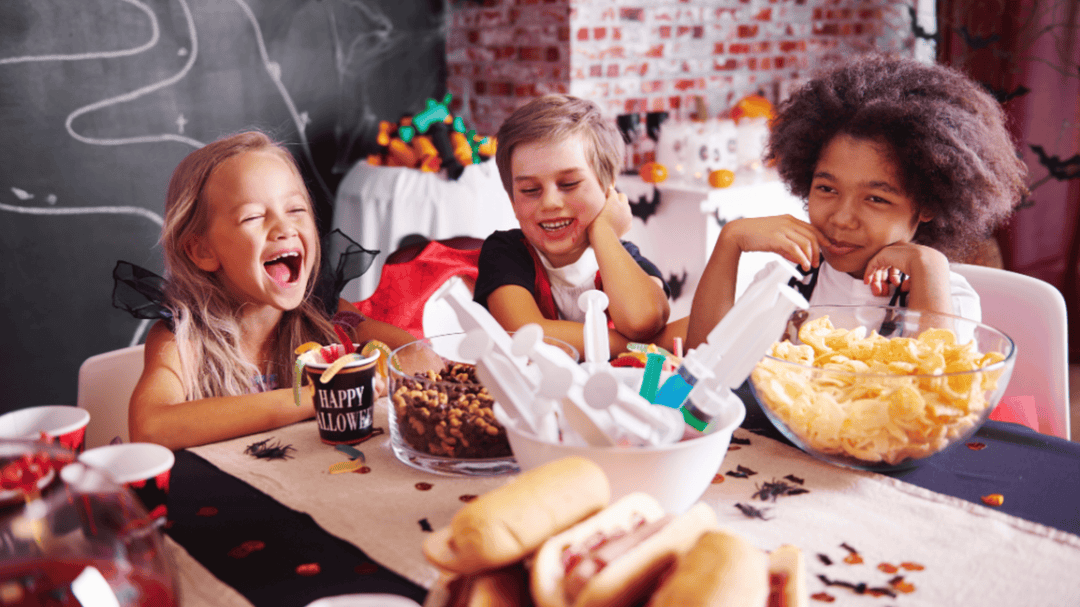 Three tweens laugh at a Halloween-themed party table with snacks, syringes, and spooky decorations.