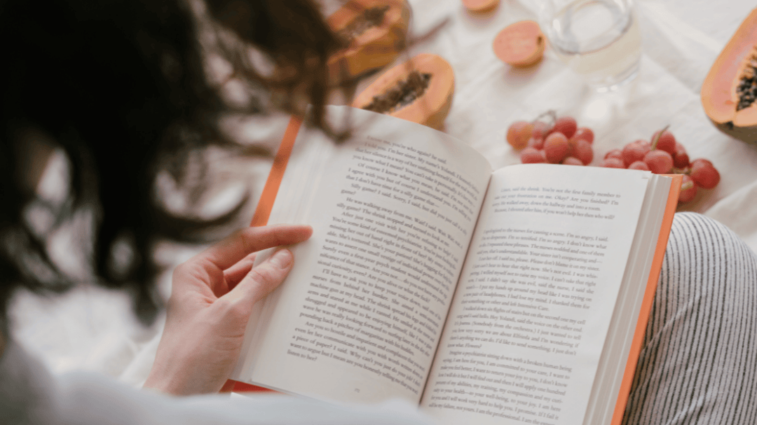 Person reading a book on a cozy blanket with papaya slices, grapes, and a drink arranged around them, creating a relaxed and inviting reading atmosphere.