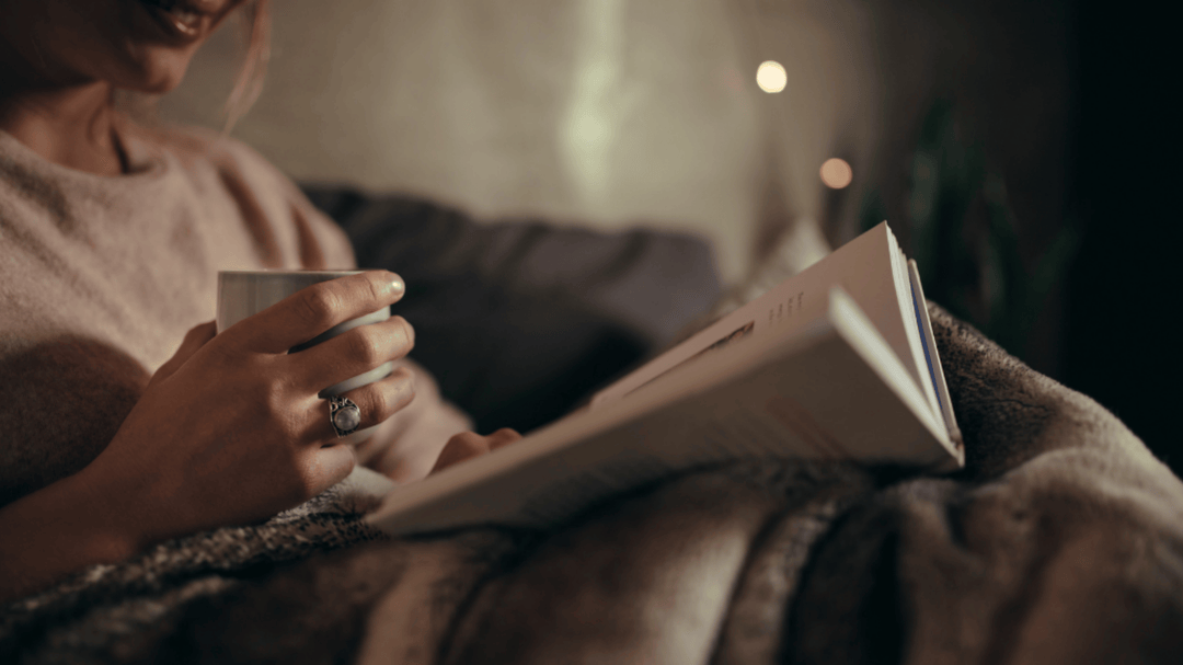 A woman in a cozy sweater enjoys a peaceful moment reading in bed, holding a warm drink, and wrapped in a soft blanket.