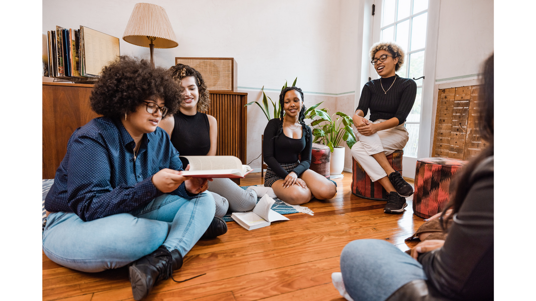 A group of young adults engaging in a lively book club discussion in a cozy room, with one reading aloud and others listening.