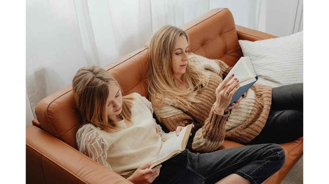 Two women, possibly a mother and daughter, comfortably reading books while reclining together on a brown leather couch, enjoying a quiet moment.