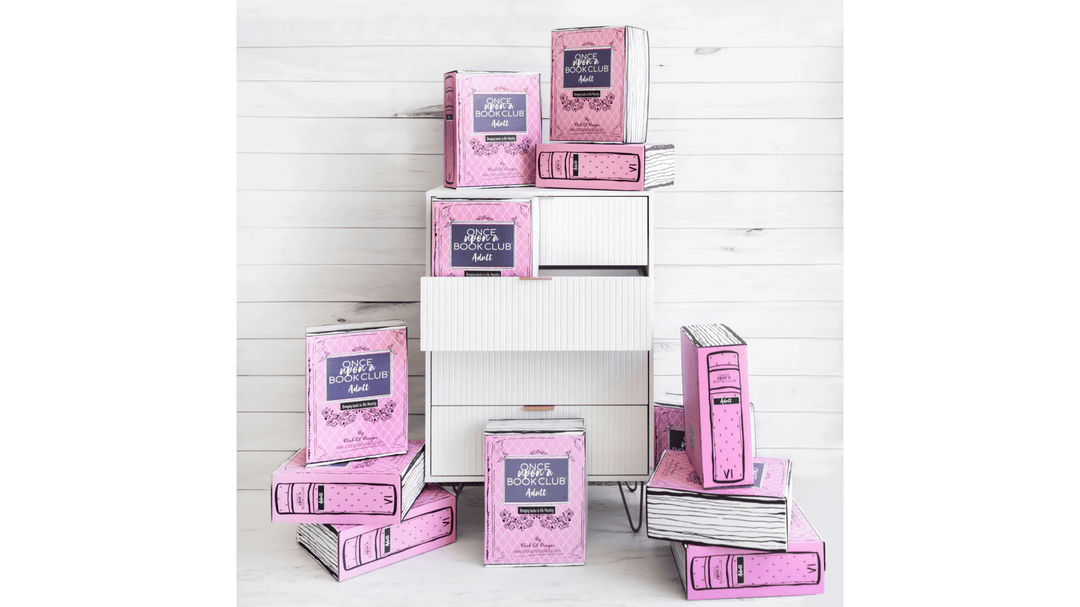 Stack of pink Once Upon a Book Club Adult boxes styled like books against a white wooden background.