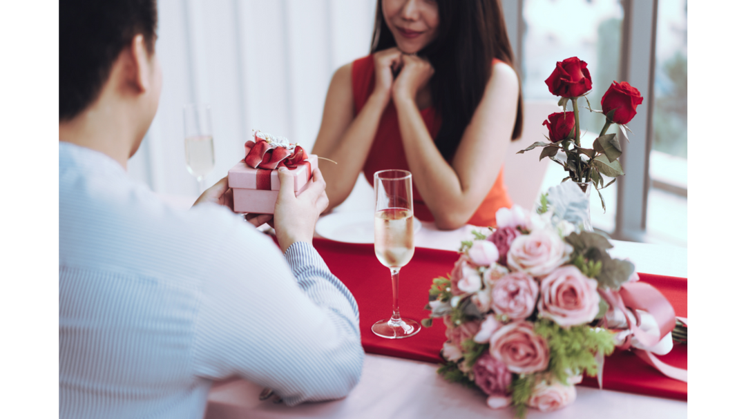 Couple at a Valentine’s Day dinner as one partner presents a small pink gift box, with champagne glasses and roses on the table.