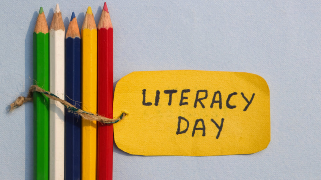 Colored pencils tied together with string next to a yellow cutout that reads “Literacy Day” on a blue background.