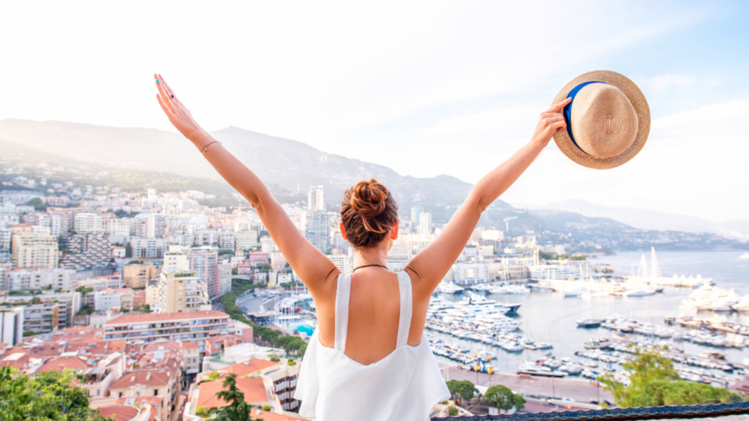 Woman overlooking a coastal city with arms raised, holding a hat, capturing a carefree travel moment.