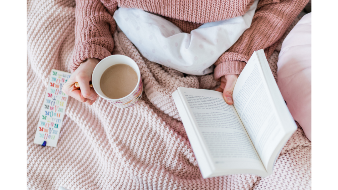 Person relaxing under a pink knitted blanket, holding a mug of coffee and reading an open book.