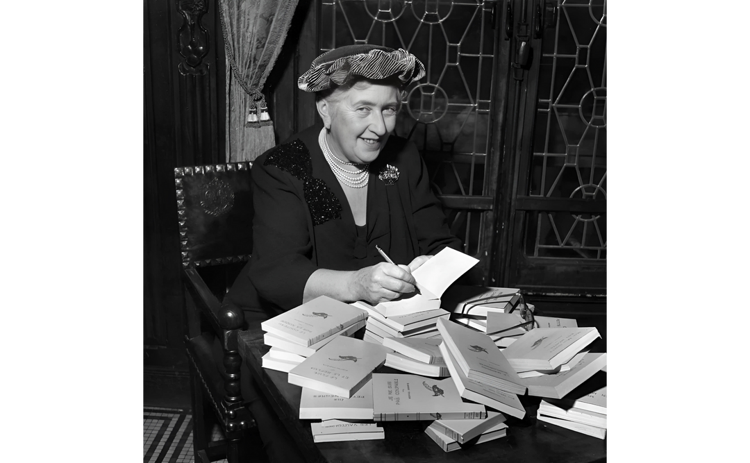 A black-and-white photo of Agatha Christie signing books at a table, surrounded by stacks of copies in an elegant vintage setting.
