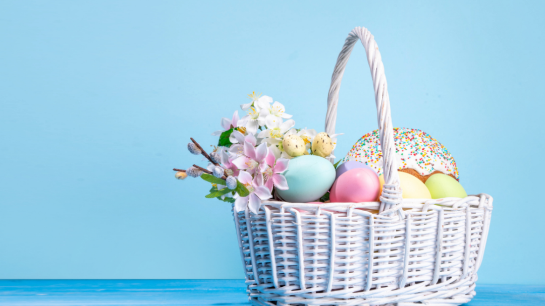 White woven Easter basket filled with pastel eggs, flowers, and a sprinkled donut, set against a soft blue background.