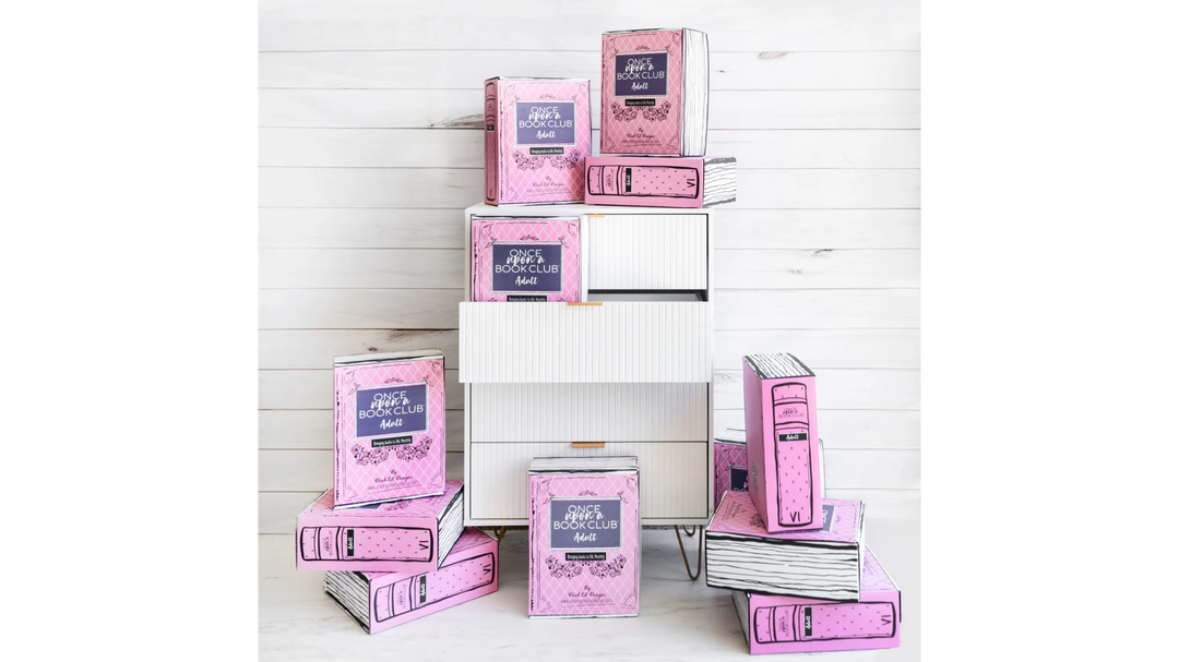 Stack of pink Once Upon a Book Club Adult subscription boxes arranged around a white modern drawer unit against a white shiplap background.