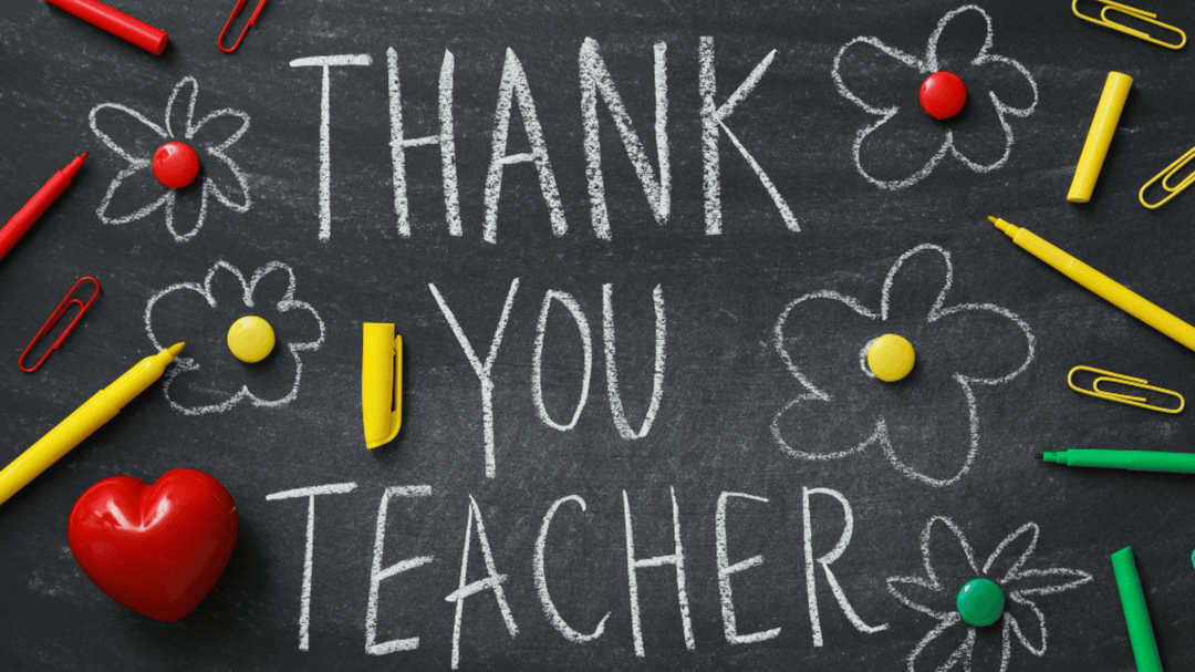 Thank you teacher written on a chalkboard with flowers, pens, clips, and a red heart placed decoratively around it.
