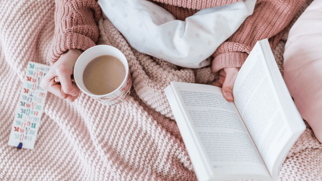 A person in a cozy pink sweater sits on a bed with a blush pink knitted blanket, holding a book in one hand and a cup of coffee in the other, with a colorful bookmark nearby.