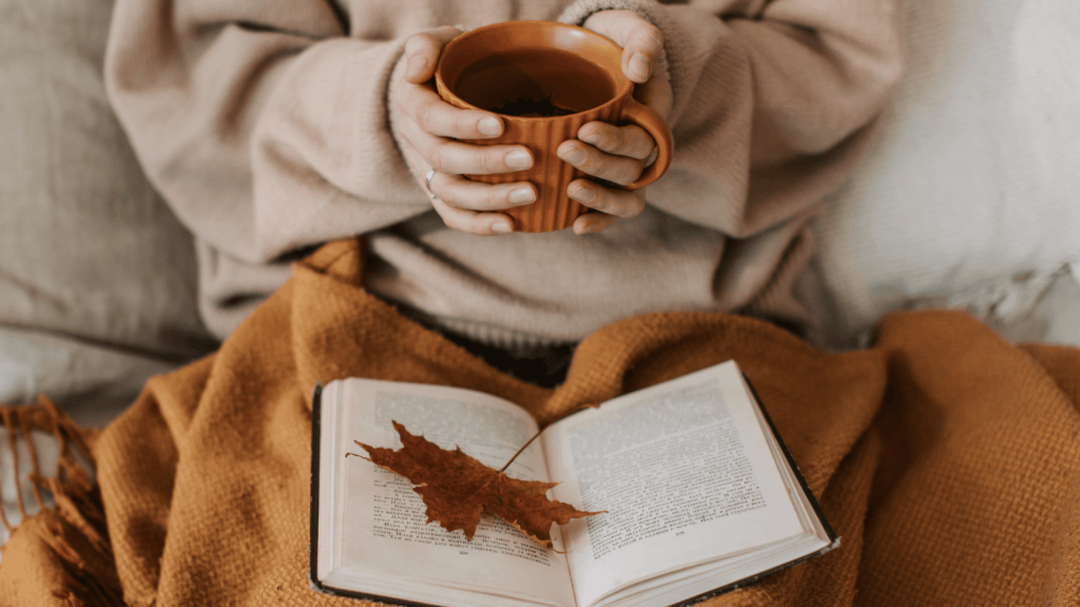 Person holding a warm mug while sitting with an open book and autumn leaf on their lap, wrapped in a cozy brown blanket.