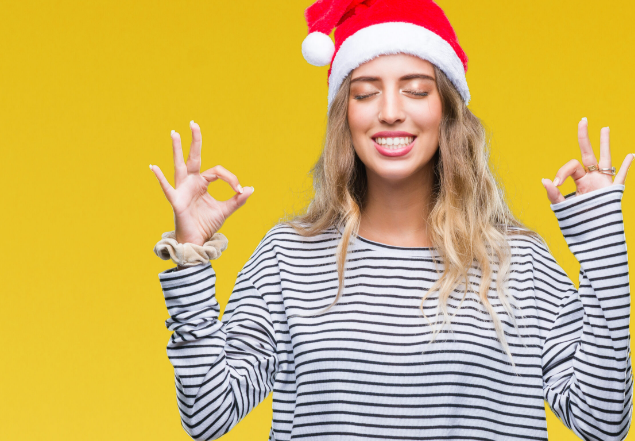 Young woman wearing a Santa hat and a striped shirt, eyes closed, and hands making an 'okay' gesture, set against a bright yellow background.