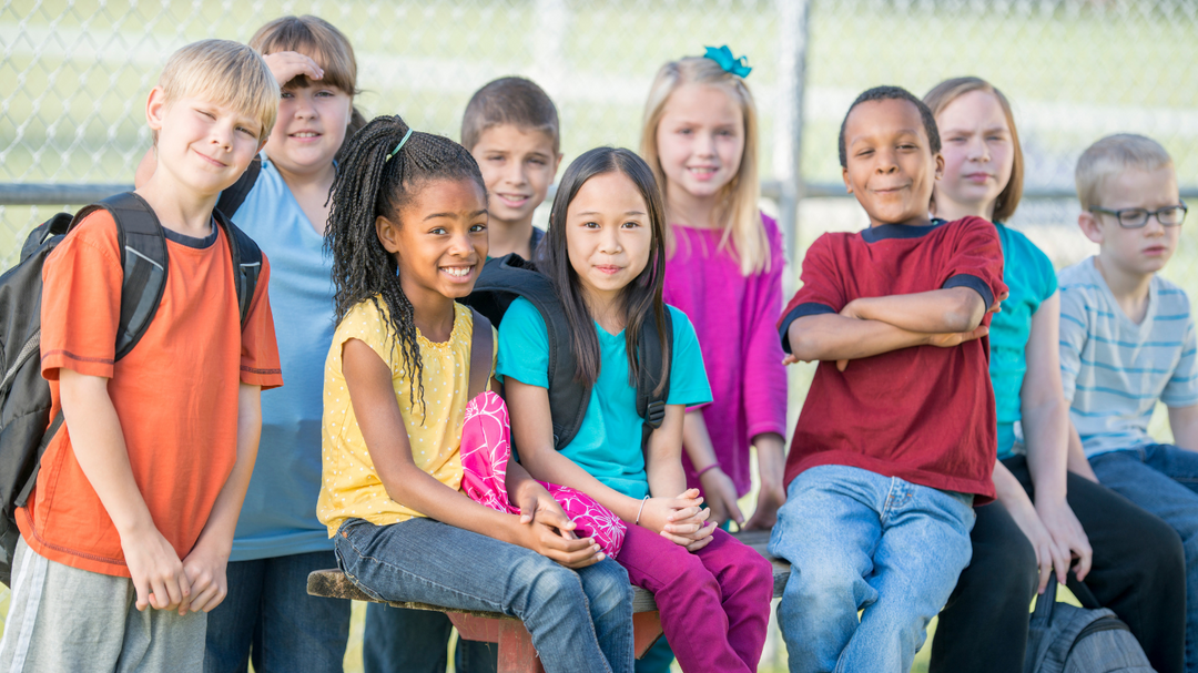 A group of diverse elementary school children smiling and sitting together on a bench outside near a chain-link fence.