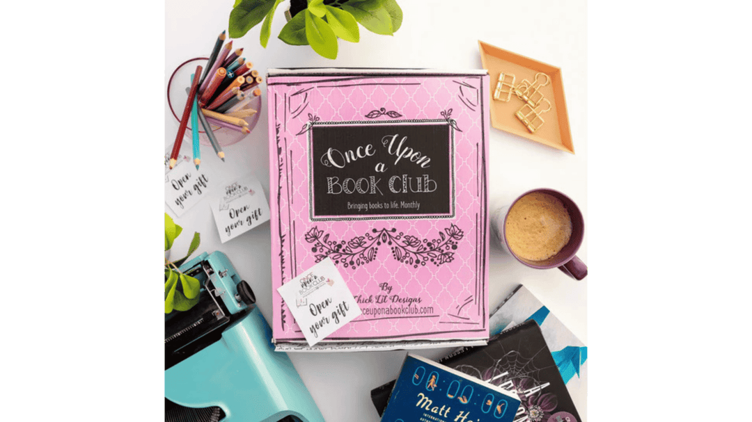 Pink Once Upon a Book Club box on a desk surrounded by books, coffee, stationery, and a typewriter.