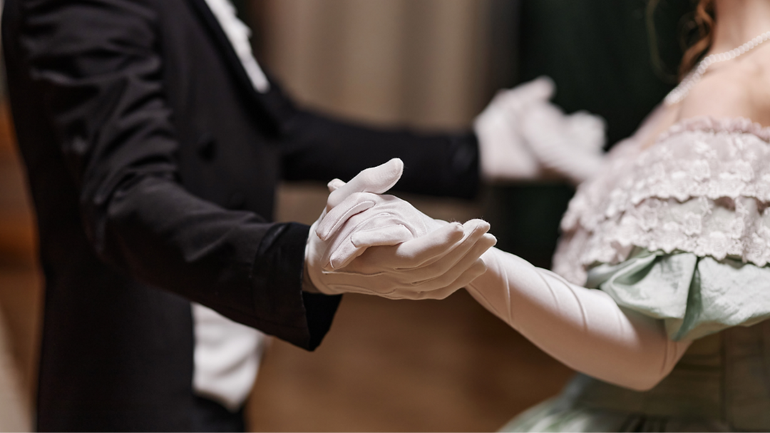 A close-up of two individuals dancing at a formal event, dressed in elegant attire, holding hands while wearing white gloves.