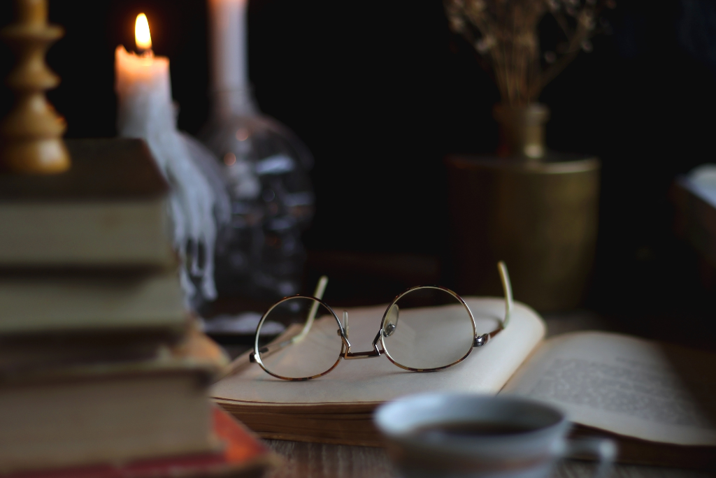 Dark academia reading scene with stacked books, an open book, round glasses, a candle, and a teacup.