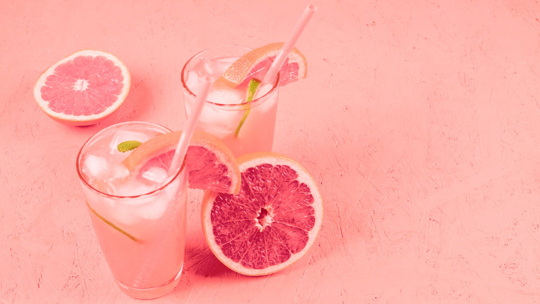 Two glasses of grapefruit lemonade with ice and pink straws on a pink background, surrounded by fresh grapefruit slices.