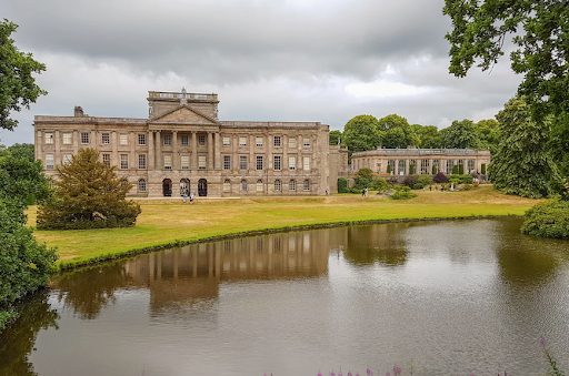 A large, historic building with manicured grounds reflected in a pond, under a cloudy sky, showcasing an elegant estate.