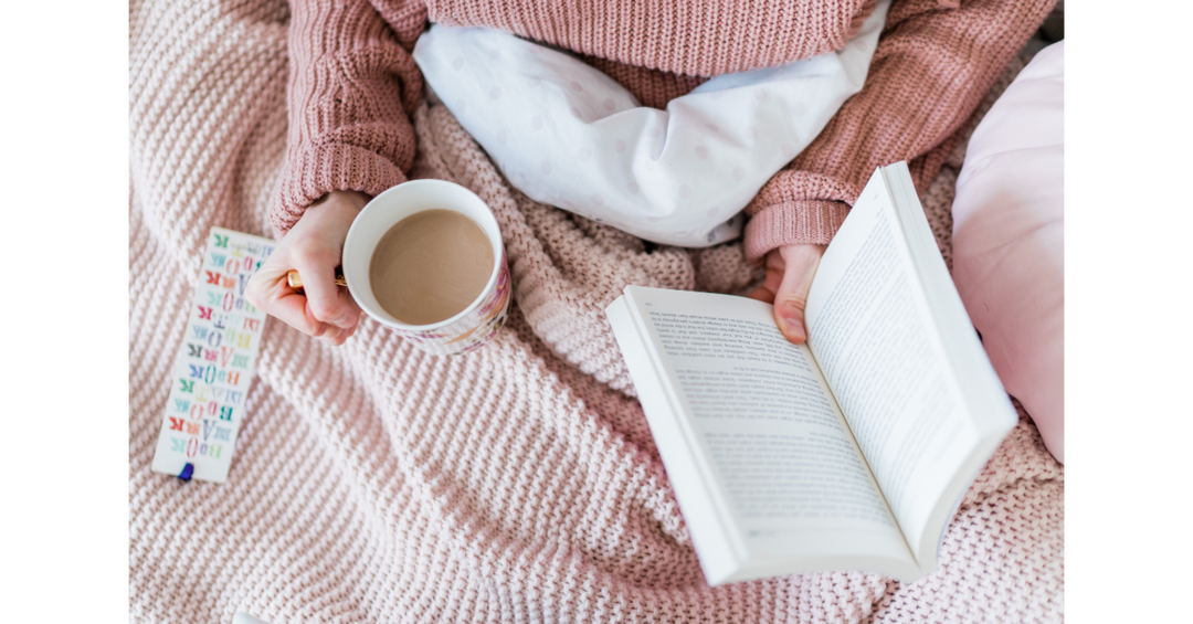 Person relaxing under a pink knitted blanket, holding a mug of coffee and reading an open book.