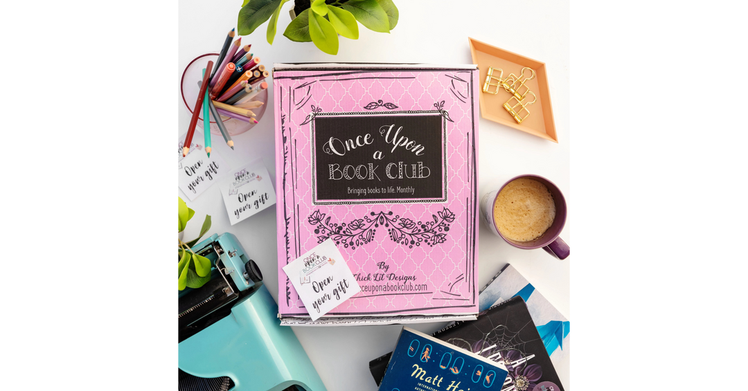 Pink Once Upon a Book Club box on a desk surrounded by books, coffee, stationery, and a typewriter.