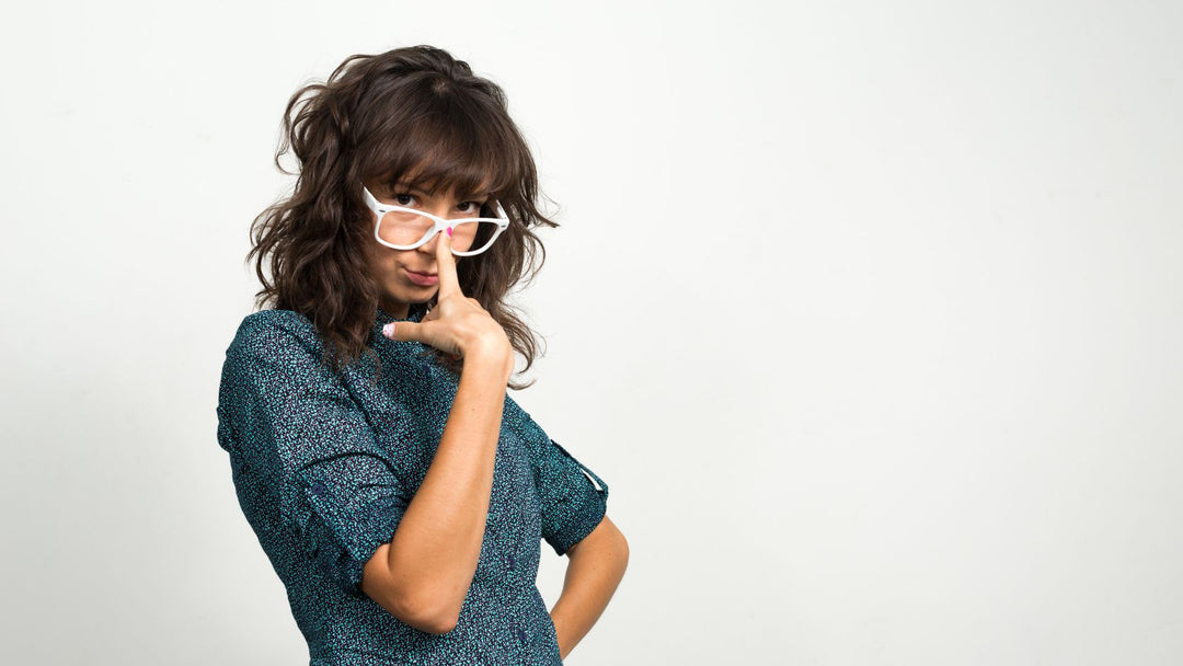 A girl with a blue dress and shoulder-length dark brown hair is lifting her white glasses.