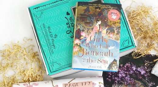 "Flat lay of 'The Girl Who Fell Beneath the Sea' book by Axie Oh, surrounded by a themed book club kit including bookmarks, a page tracker, and decorative items."