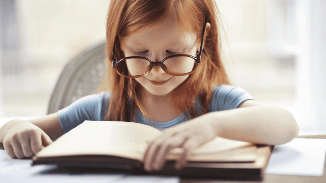 Young red-haired girl with glasses reading a book at a desk with focused expression.