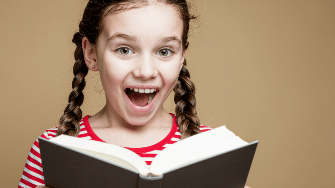 Excited girl in pigtails reading a book with wide eyes and a big smile against a plain tan background.