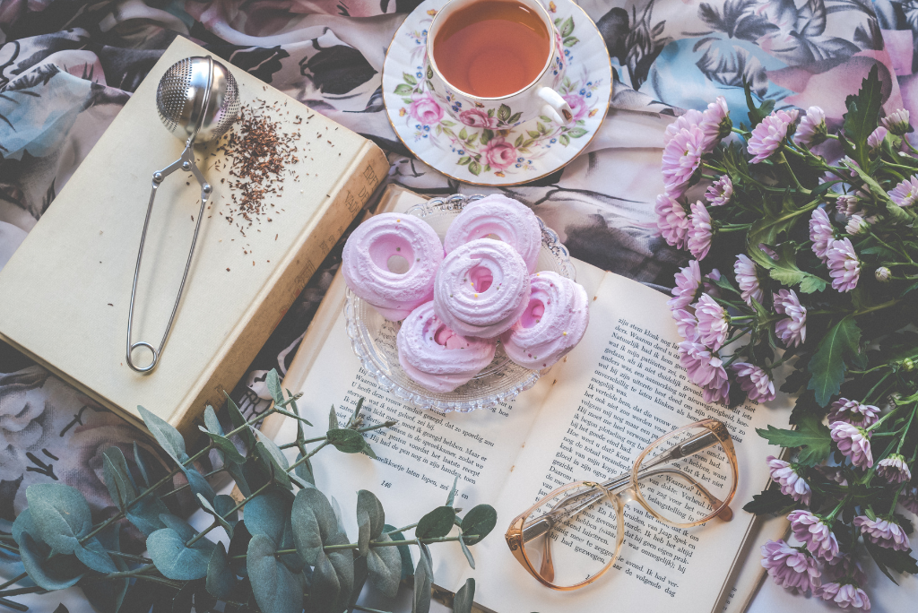 A cozy scene with an open book, reading glasses, pink macarons, a tea strainer, and a teacup, styled with fresh flowers for a relaxing reading experience.