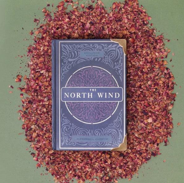 The book 'The North Wind,' written by Alexandria Warwick, is placed atop a bed of dried red rose petals.