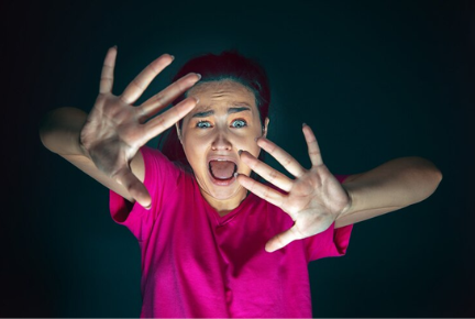 A woman wearing a pink shirt appears terrified, with wide eyes and hands raised defensively, as if reacting to something frightening in the dark.