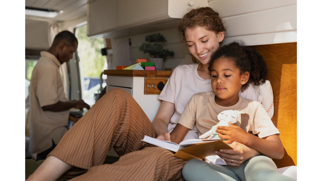 A mother and daughter sit together reading a book in a cozy space, while a man prepares food in the background, creating a warm atmosphere.