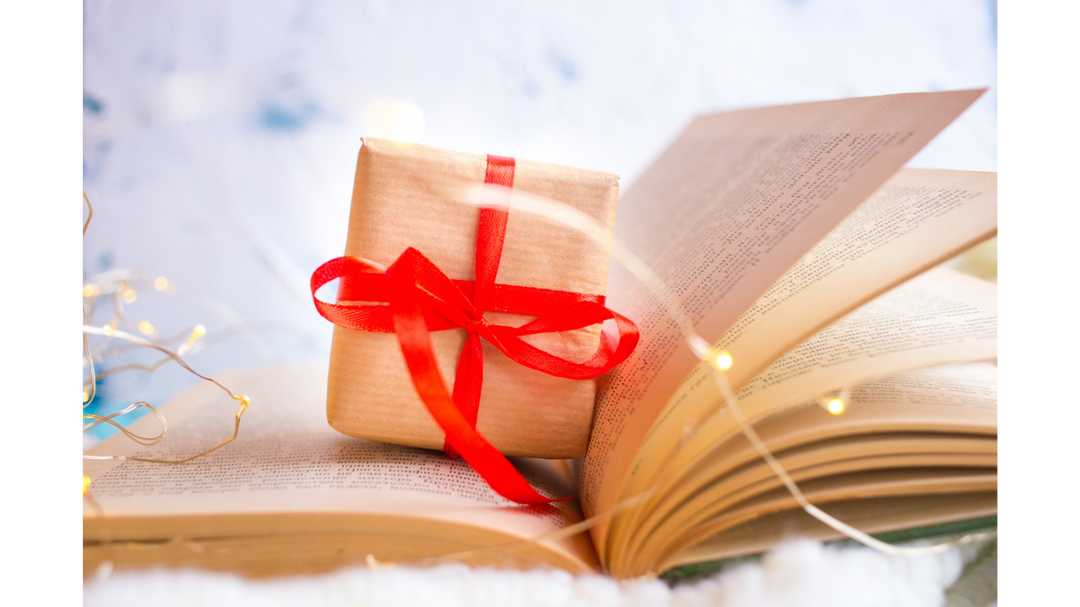 A small gift wrapped in brown paper with a red ribbon sits on an open book, surrounded by fairy lights, evoking a cozy reading atmosphere.