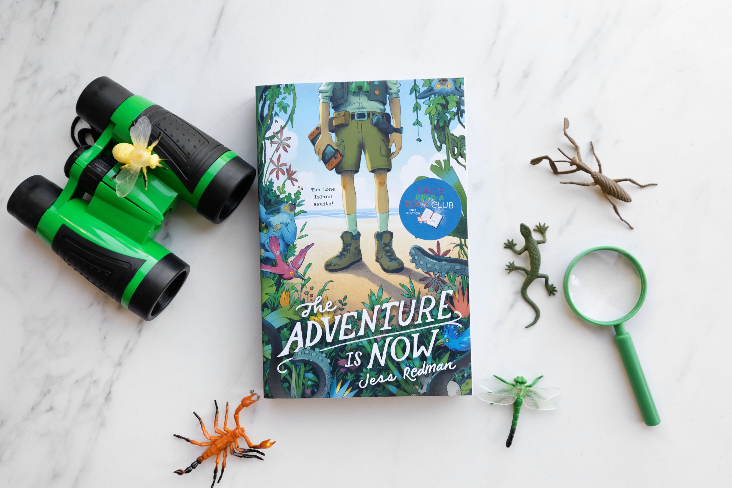 A collection of some of the items from The Brave Explorer Middle Grade box from August 2023. These include the featured book, The Adventure is Now by Jess Redman, a collection of fake rubber insects, a magnifying glass, and a pair of binoculars.
