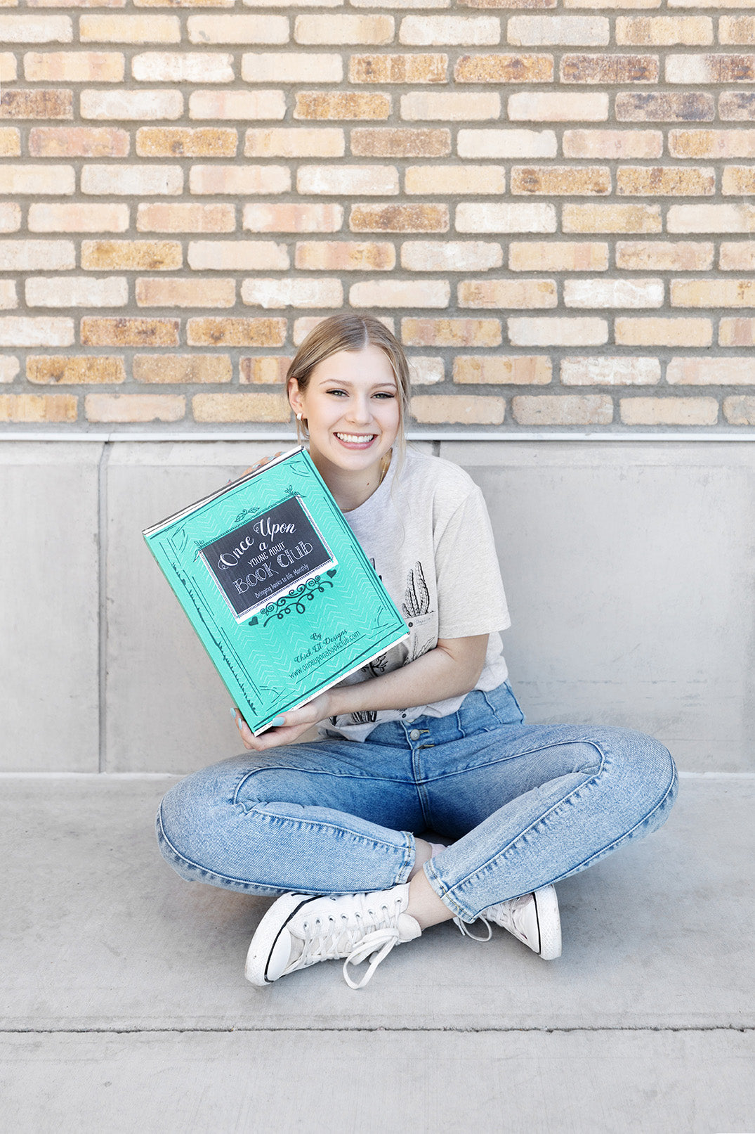 a white blonde woman wearing a beige shirt, blue jeans, and white converse sits on the ground in front of a brick wall holding a green Once Upon a Book Club box and smiling
