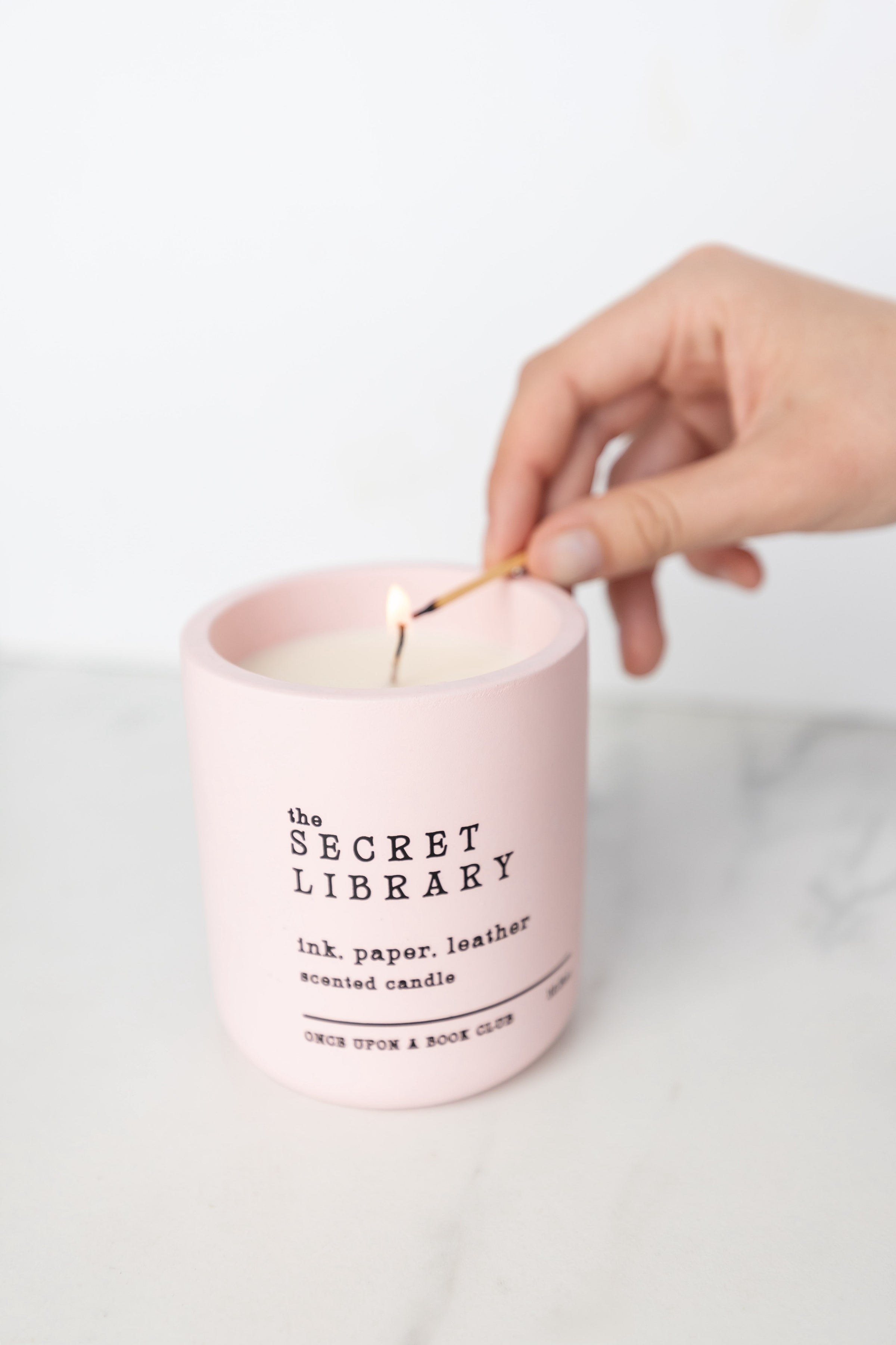 A white hand holds a match to a ceramic pink candle. The words The Secret Library are written in larger letters. Under that are the words ink. paper. leather. scented candle. Once Upon a Book Club.