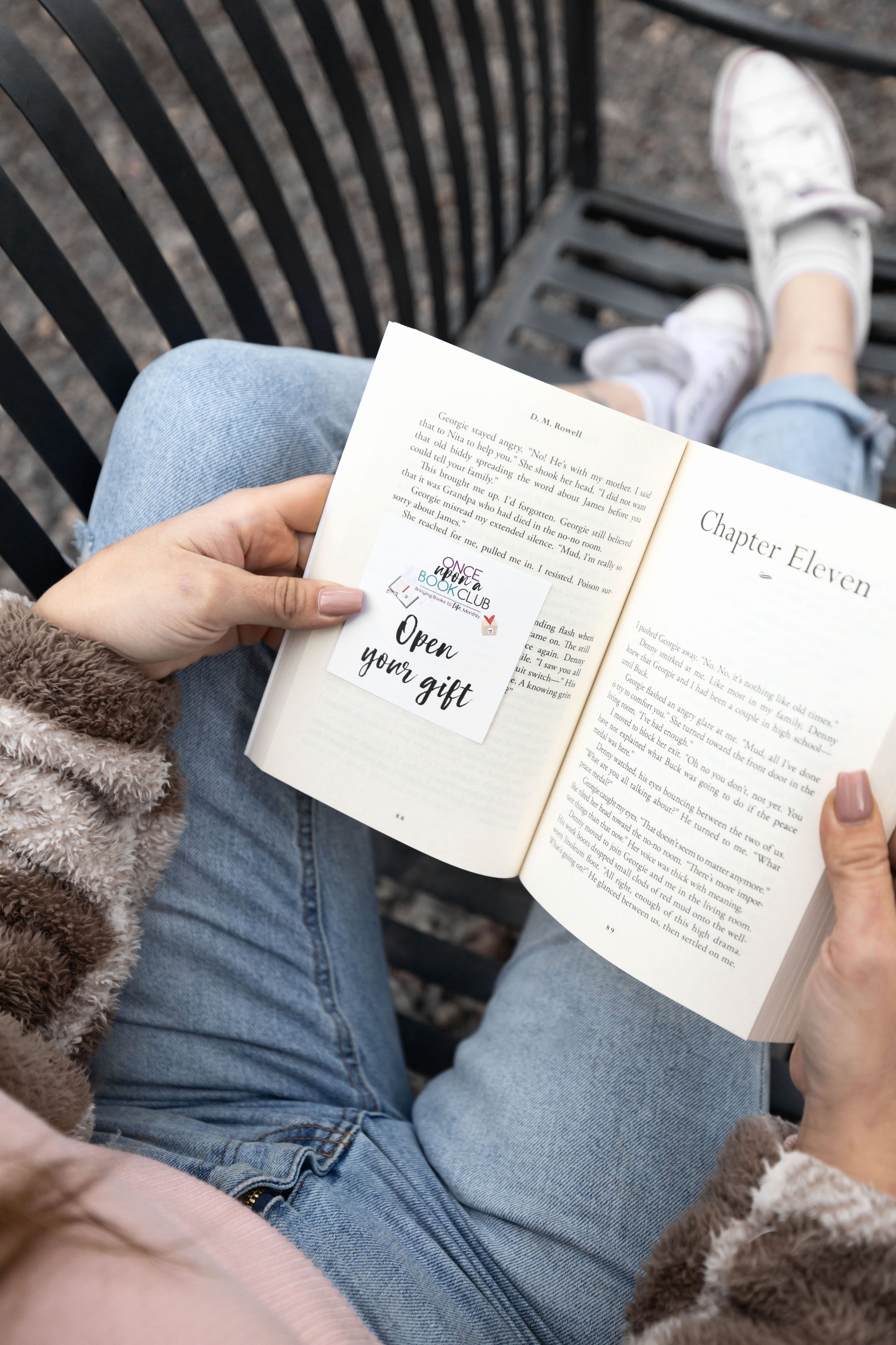 A pair of white hands holds open a book on a bench. Inside the book is a sticky note showing Open Your Gift to signify the reader should open the corresponding gift that is mentioned at that moment in the story.