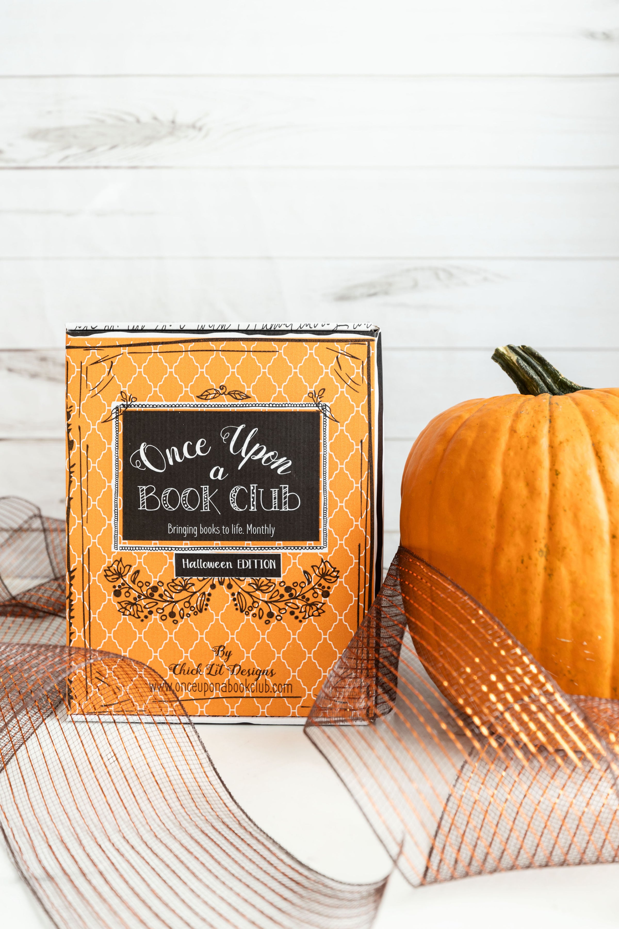 An orange Once Upon a Book Club Halloween Edition box sits next to a pumpkin. Bronze ribbon surrounds the two.