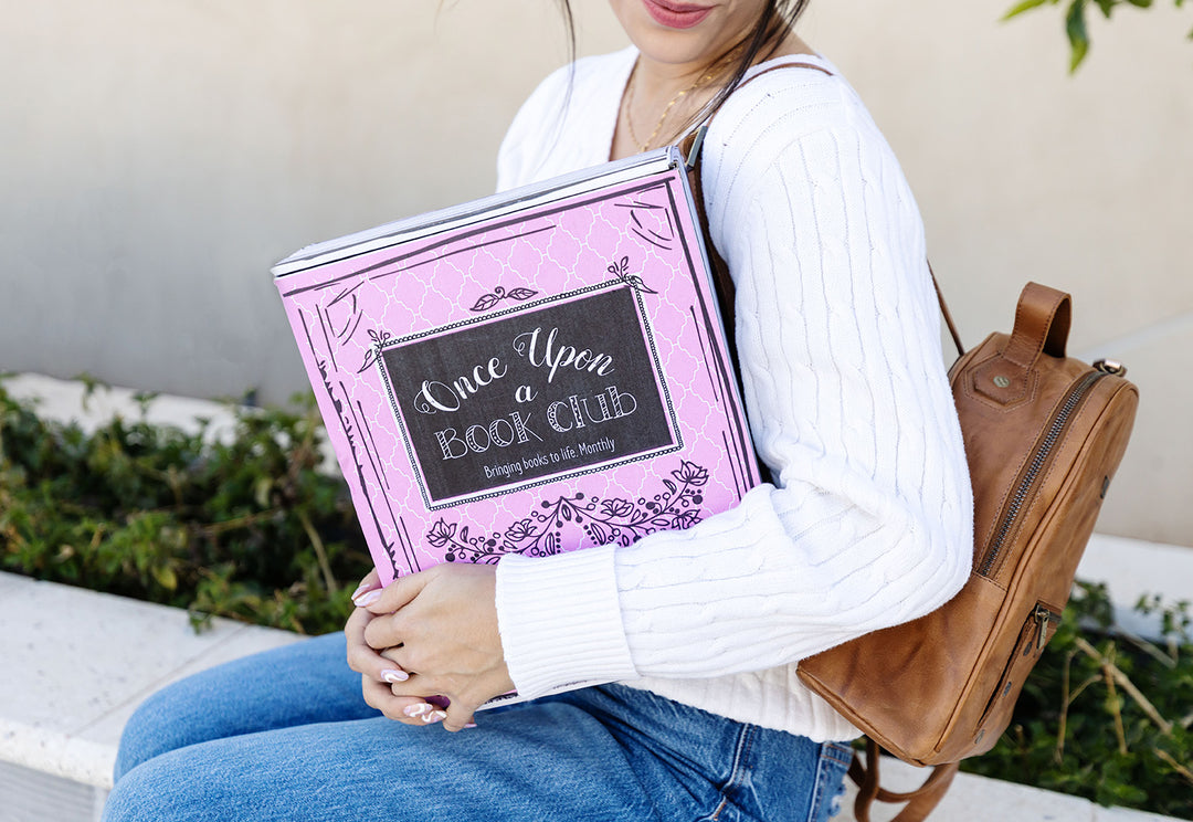 a white woman wearing a white sweater and blue jeans with a brown backpack holds a pink Once Upon a Book Club box to her side