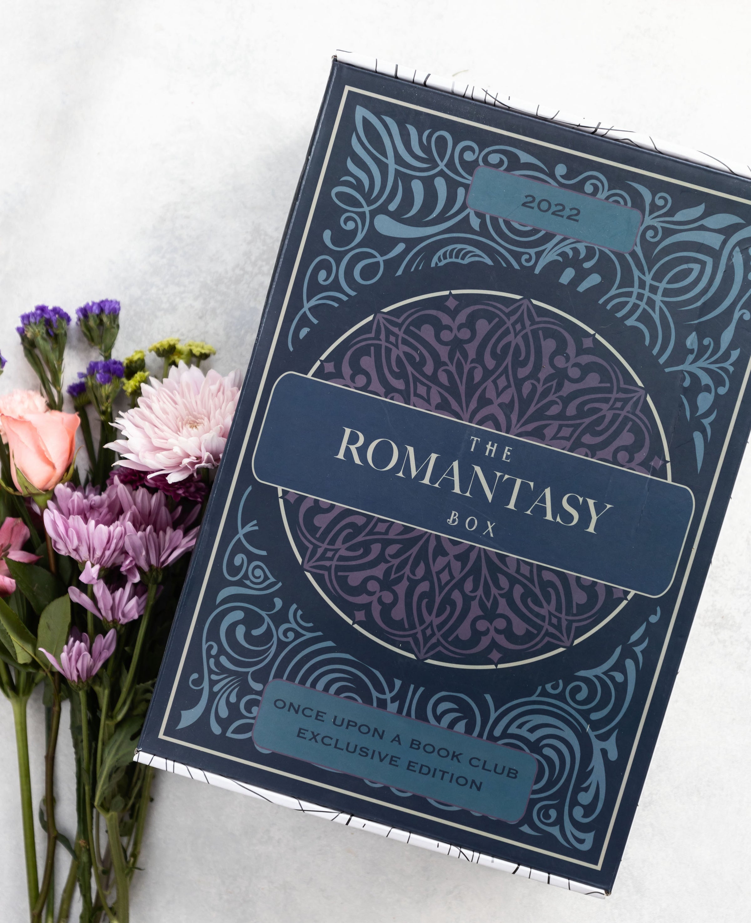 A large blue and purple special edition box titled The Romantasy Box sits on a white surface. Purple and pink flowers lay in a bouquet next to the box.