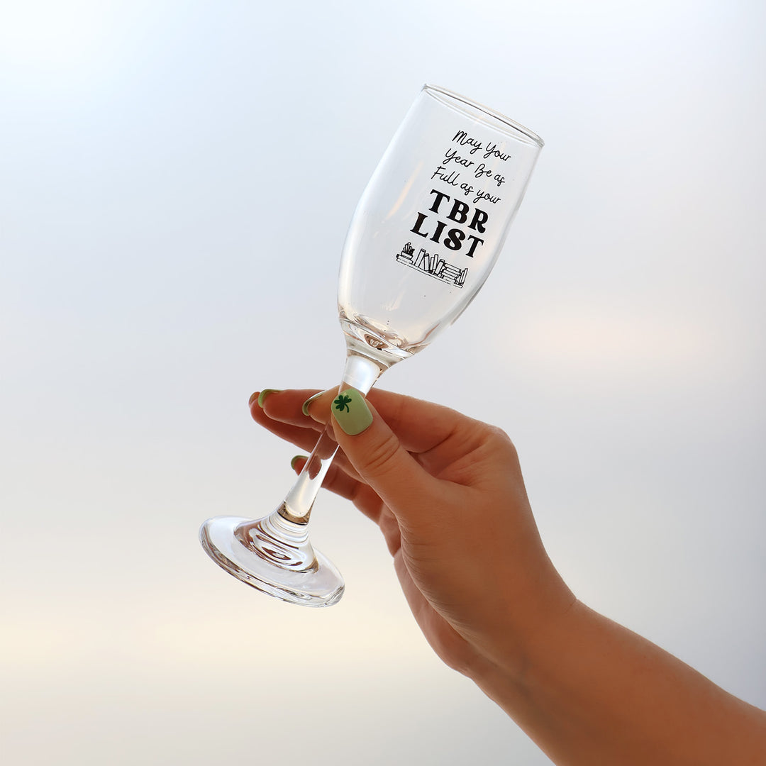 Hand holding a champagne flute that reads "May your year be as full as your TBR List" and features an etching of books along the bottom of the glass.