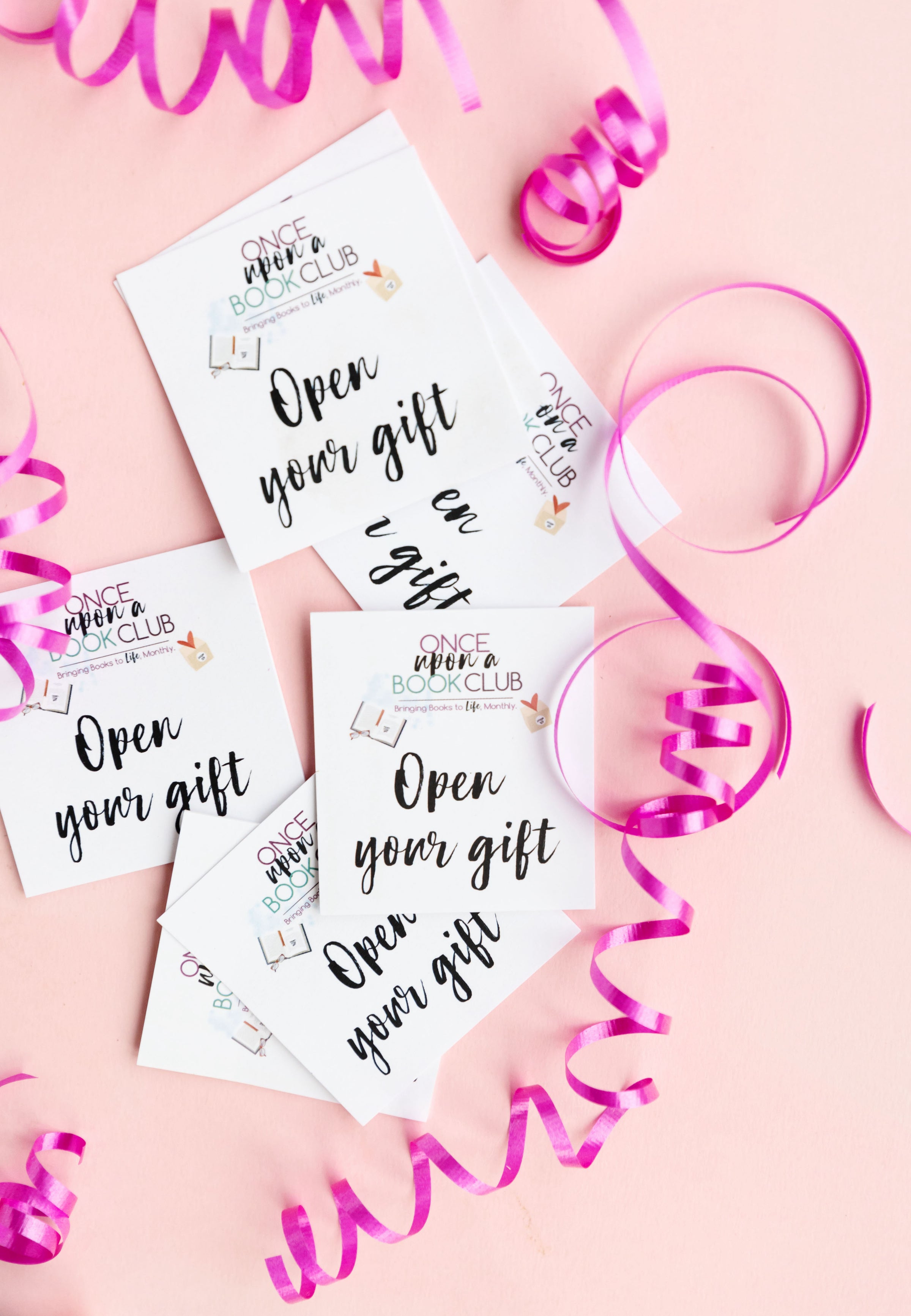 A collection of Open Your Gift sticky notes haphazardly laying on a pink background surrounded by hot pink ribbon.